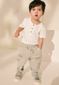 Světle béžová krátký rukáv henley košile s hnědými knoflíky, doplněná světle šedými kalhotami s hravým vzorem usměvavých obličejů a šedými teniskami.