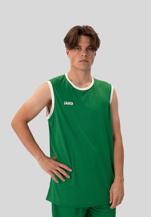 Młody mężczyzna w zielonej bezrękawnikowej koszulce sportowej i krótkich spodenkach stoi z jedną ręką na biodrze na gładkim jasnoszarym tle.