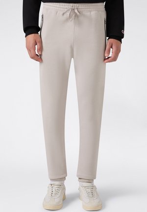 Hellbeige Sweatpants mit elastischem Bund, weiße Schnürsneaker, schwarzes Langarmshirt mit Logo auf dem Ärmel, Hände locker an den Seiten.