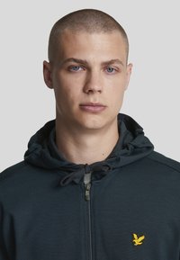 Dunkelmarineblaues Zip-Hoodie mit strukturiertem Finish, ausgestattet mit einer Kordelzugkapuze und einem kleinen gelben Vogel-Logo auf der Brust.