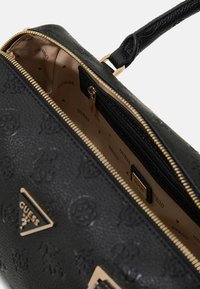 Borsa nera in materiale sintetico con dettagli dorati, chiusura con zip e esterno decorato. Interno foderato beige con logo "GUESS".