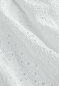 Tissu blanc texturé avec un motif de jour légèrement visible, comprenant des découpes en forme de diamant et arrondies, idéal pour des vêtements légers ou de la décoration.