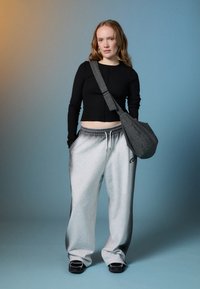 Junge Frau mit langen roten Haaren, die ein schwarzes Langarmoberteil, graue Sweathosen, schwarze Sneaker und eine karierte Umhängetasche auf einem Farbverlaufs-Hintergrund trägt.