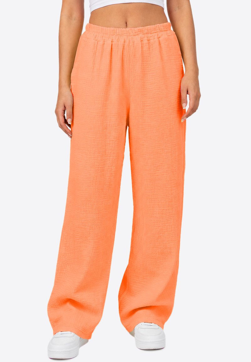 Femme portant un pantalon large, ample, texturé, orange vif avec une taille élastique et des baskets blanches, montrée de la taille aux pieds.