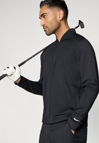 Άνδρας με μαύρο μπλουζάκι μακρύ μανίκι και γάντι γκολφ, κρατώντας ένα μπαστούνι γκολφ ακουμπισμένο στον ώμο του, κοιτάζει στο πλάι σε ανοιχτό φόντο.