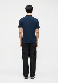 Άνδρας που στέκεται με την πλάτη, φορώντας μπλε σκούρο πόλο πουκάμισο, μαύρο παντελόνι cargo και λευκά αθλητικά παπούτσια σε απλό λευκό φόντο.