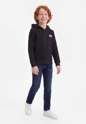 Garçon roux portant un sweat à capuche noir zippé, un jean bleu et des baskets blanches, souriant en se tenant debout sur un fond blanc uni.