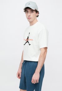 Νέος άνδρας που φοράει άσπρο t-shirt Air Jordan, μπλε σορτς και άσπρο καπέλο με λογότυπο μπάσκετ, στέκεται μπροστά σε μια απλή φόντο.