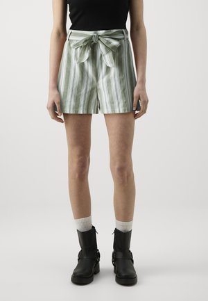 Persona con pantalones cortos a rayas verdes y blancas con un cinturón atado, botas negras, calcetines blancos y una camiseta sin mangas negra.