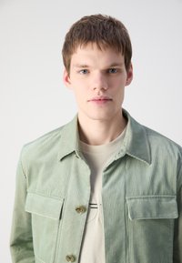 Jachetă din corduroy verde deschis, cu două buzunare pe piept, închidere cu nasturi și un finisaj texturat, purtată peste un tricou bej.