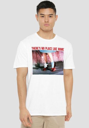 Young man wearing white T-shirt with "THERE'S NO PLACE LIKE HOME" text and image of legs in red shoes and blue socks holding a wand.