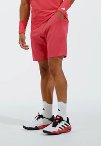 Shorts de sport rouge avec poches latérales, associés à des chaussures blanches et noires avec des accents rouges et des semelles noires. Chaussettes de sport blanches visibles.