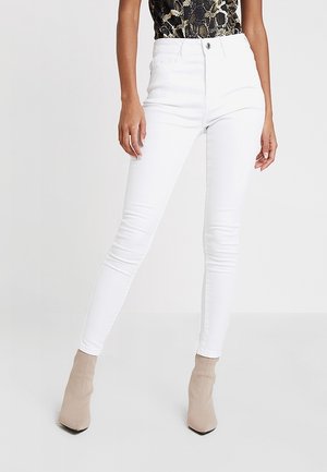 Kvinde iført højtaljede hvide skinny jeans, top med slangeskindsmønster og beige ankelstøvler med spids tå, stående mod en ensartet hvid baggrund.