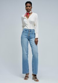 Mujer vestida con blusa blanca, jeans de tiro alto azul claro con botones, bufanda roja alrededor del cuello, pendientes de aro dorados y sandalias de tacón blancas.