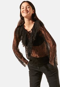Blouse en dentelle noire avec un col volanté et des manches longues, dotée d'une fermeture à nouer au col et d'une texture transparente, superposée à un vêtement sombre.