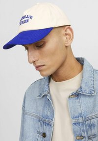 Baseball-cap i creme og blå med broderet tekst. Båret med en denimjakke over en beige skjorte, der fremhæver et afslappet design.