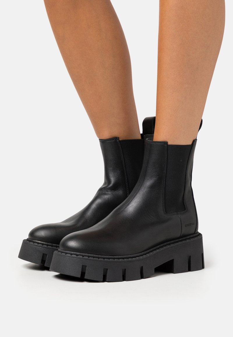 Bottines en cuir noir avec semelles épaisses et rainurées portées sur des jambes nues sur un fond clair uni.
