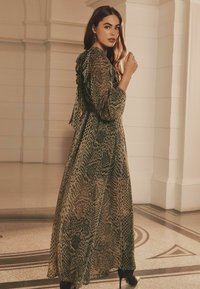 Rochie lungă, fluidă, fabricată dintr-un material ușor, cu imprimeu de șarpe în tonuri de verde și bej, având un decolteu volanat și mâneci lungi. Pantofi cu toc negri.