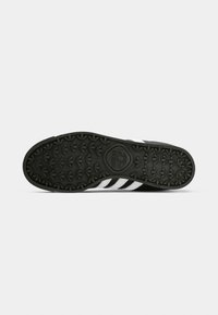 Fekete Adidas cipőtalp texturált trefoil logómintával és oldalt látható fehér csíkokkal, egyszerű fehér háttér előtt.