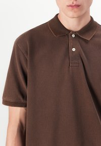 Brown cotton polo shirt with a textured fabric, short sleeves, and a collar featuring three button closures.