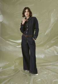 Combinaison en denim bleu foncé avec des coutures dorées, ajustée en haut et à pattes larges. Comprend des fermetures à boutons et une ceinture marron.