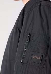 Chaqueta bomber negra con un tejido texturizado y arrugado. Incluye un bolsillo con cremallera en la manga izquierda y un parche de logo en tela.