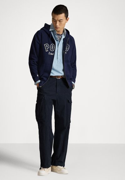 Moški v mornarsko modri jopiči z zadrgo Polo, svetlo modri srajci z gumbi čez črtasto majico, temnih hlačah s sedeži in svetlo bež supergah, stoji z rokami v žepih.