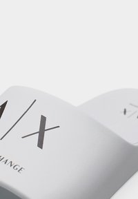 Κοντινή λήψη μιας καμπυλωτής λευκής ετικέτας με μαύρο κείμενο "A/X" και "EXCHANGE" σε απλό ανοιχτόφωτο φόντο.