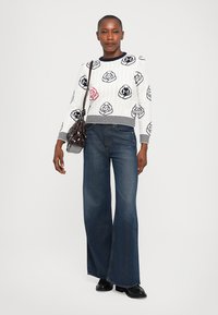 Weißer Strickpullover mit schwarzen und roten Rosenmustern, kombiniert mit hochtaillierten, weit geschnittenen blauen Jeans. Das Model hält eine kleine braune Tasche.