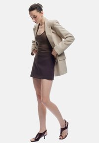 Beige blazer over een bruine tanktop en een donkerbruine minirok. Zwarte open teen sandalen met hakken. Soepele texturen met een aansluitende silhouet.