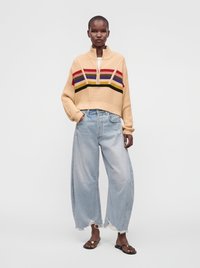 Personne portant un pull court beige à rayures colorées, un jean large bleu clair déchiré et des sandales noires avec un détail doré.
