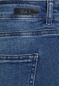 Tecido de denim em lavagem azul escura, com um patch na cintura com "NOOILY MAY" em letras pretas em relevo e um design de bolso traseiro.