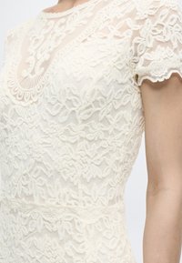 Záber zblízka osoby nosiacej biely krajkový šaty s krátkym rukávom, kvetinovými vzormi a zdobeným výstrihom s vlnitým okrajom.