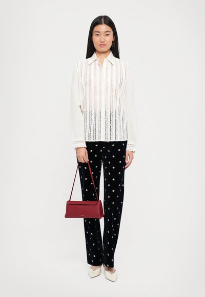 Naistele, kes kannab valget pitsist pikakäist blouse, musti lillelise mustriga pükse, valgeid terava ninaga kingi, hoiab punast ristkülikukujulist käekoti selle rihmast.