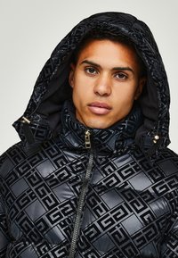 Black puffer jacket featuring glossy finish, embossed geometric pattern, high collar, and adjustable hood. Zipper and hardware accents included.