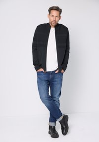 Chaqueta negra con cierre de cremallera y detalles texturizados, camiseta blanca, jeans azules y botines negros, posando frente a un fondo liso.