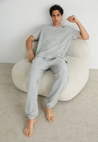 Lichtgrijze katoenen lounge-set, bestaande uit een comfortabel t-shirt met een relaxte pasvorm en joggingsbroek, zittend op een luxe, ronde crème kleurige stoel.