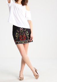 Biała bluzka na jedno ramię z koronkowym wykończeniem, w połączeniu z czarną spódnicą z kolorowym haftem w kwiaty i dekoracyjnym wykończeniem. Jasne buty.