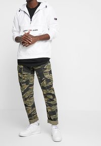 Biała kurtka przeciwdeszczowa z kieszenią z przodu i logo, połączona z zielonymi spodniami cargo w kamuflażu i białymi sneakersami. Casualowy i funkcjonalny strój.