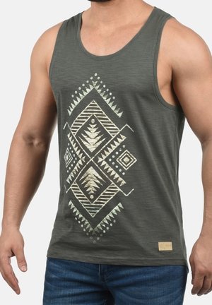 Homme portant un débardeur vert foncé sans manches avec un motif tribal géométrique beige symétrique, associé à un jean bleu.