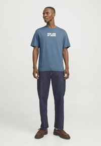 Blå bomulds T-shirt med korte ærmer og rund hals, med et hvidt "JACK & JONES" logo på forsiden. Parret med mørk denim jeans og brune sko.