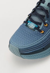 Zapatilla deportiva azul con una parte superior de malla texturizada, que cuenta con una suela gruesa y acolchada, acentos en navy contrastantes y un detalle de cordones naranjas.