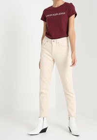 Camiseta de algodón burdeos con el texto blanco "Calvin Klein Jeans", combinada con jeans color crema y botas blancas brillantes de caña alta con tacones en cuña.