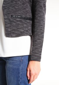 Veste grise texturée au design raccourci, avec une poche accent en cuir noir. Portée sur une chemise blanche et un jean en denim.