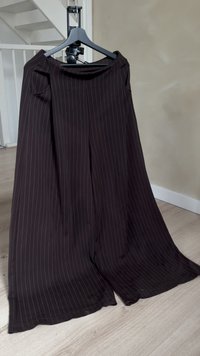 Wijd uitlopende broek van donkerbruine stof met witte verticale strepen. Beschikt over een gladde tailleband en een vloeiend ontwerp. Hangt aan een zwarte hanger.