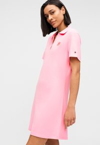 Φόρεμα πόλο από ανοιχτό ροζ ύφασμα, με γιακά και κοντά μανίκια. Κεντημένο λογότυπο στο στήθος και αντίθετο διακοσμητικό στα μανίκια.