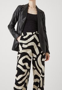 Zwarte faux leren blazer gecombineerd met beige, wijde broek met gedurfde zwarte abstracte patronen. Getextureerde top onder de blazer.