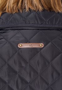 Vadderad svart jacka med diagonala sömmar, ett brunt lädermärke med guldfärgade detaljer och en slät, glänsande yta.