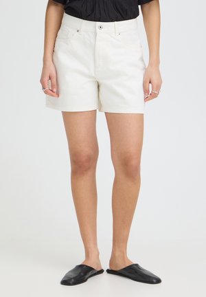 Személy fehér magasderekú rövidnadrágot, fekete felsőt, fekete bebújós cipőt és ezüst gyűrűket visel az ujjain, egyszínű háttér előtt.