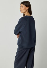 Frau mit langen dunklen Haaren, die einen locker sitzenden navy-blauen Sweatshirt und passende Hosen trägt, seitlich vor einem schlichten hellen Hintergrund stehend.